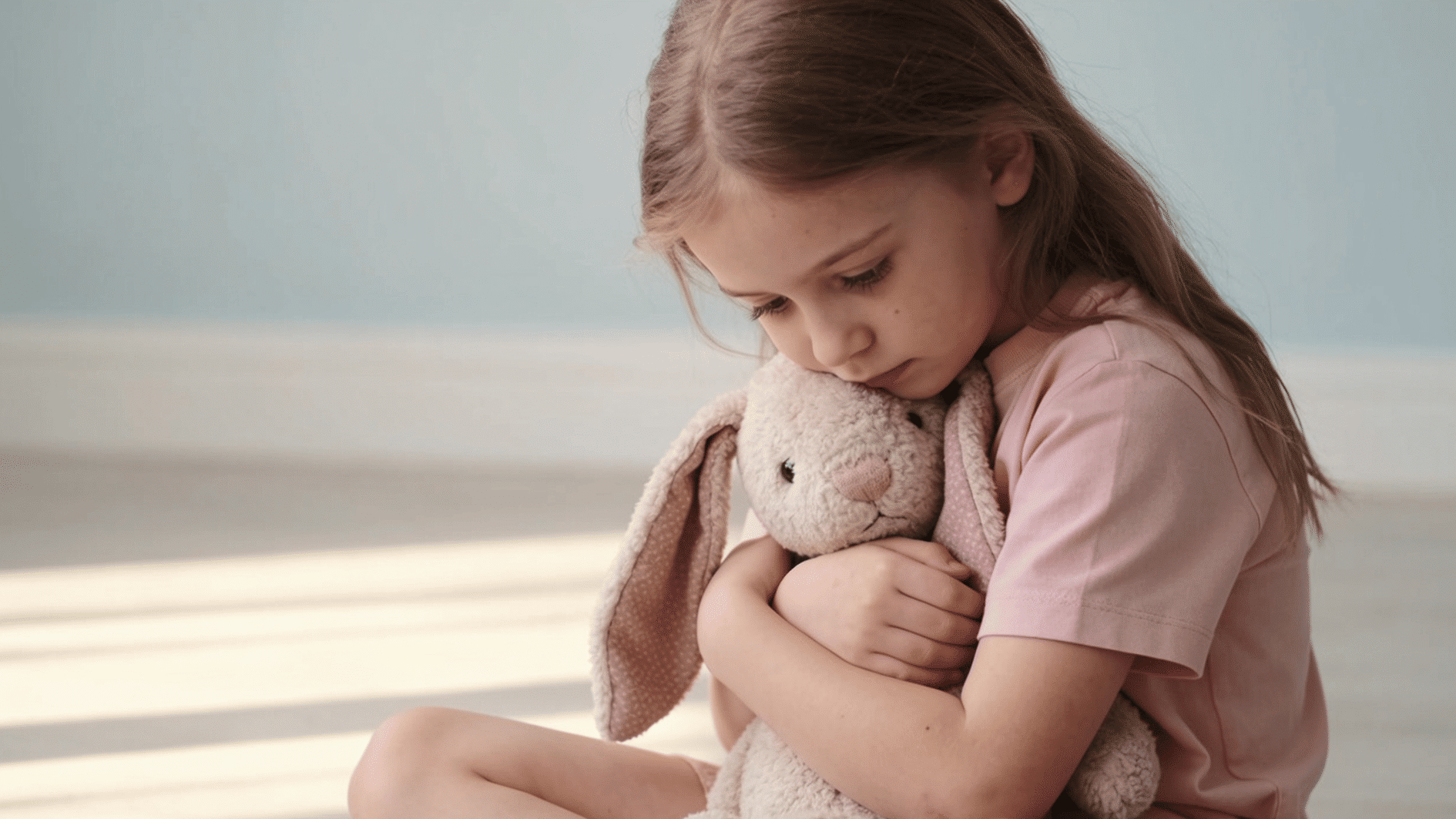 young girl sitting on the floor hugging a stuffed bunny with a withdrawn expression representing childhood trauma