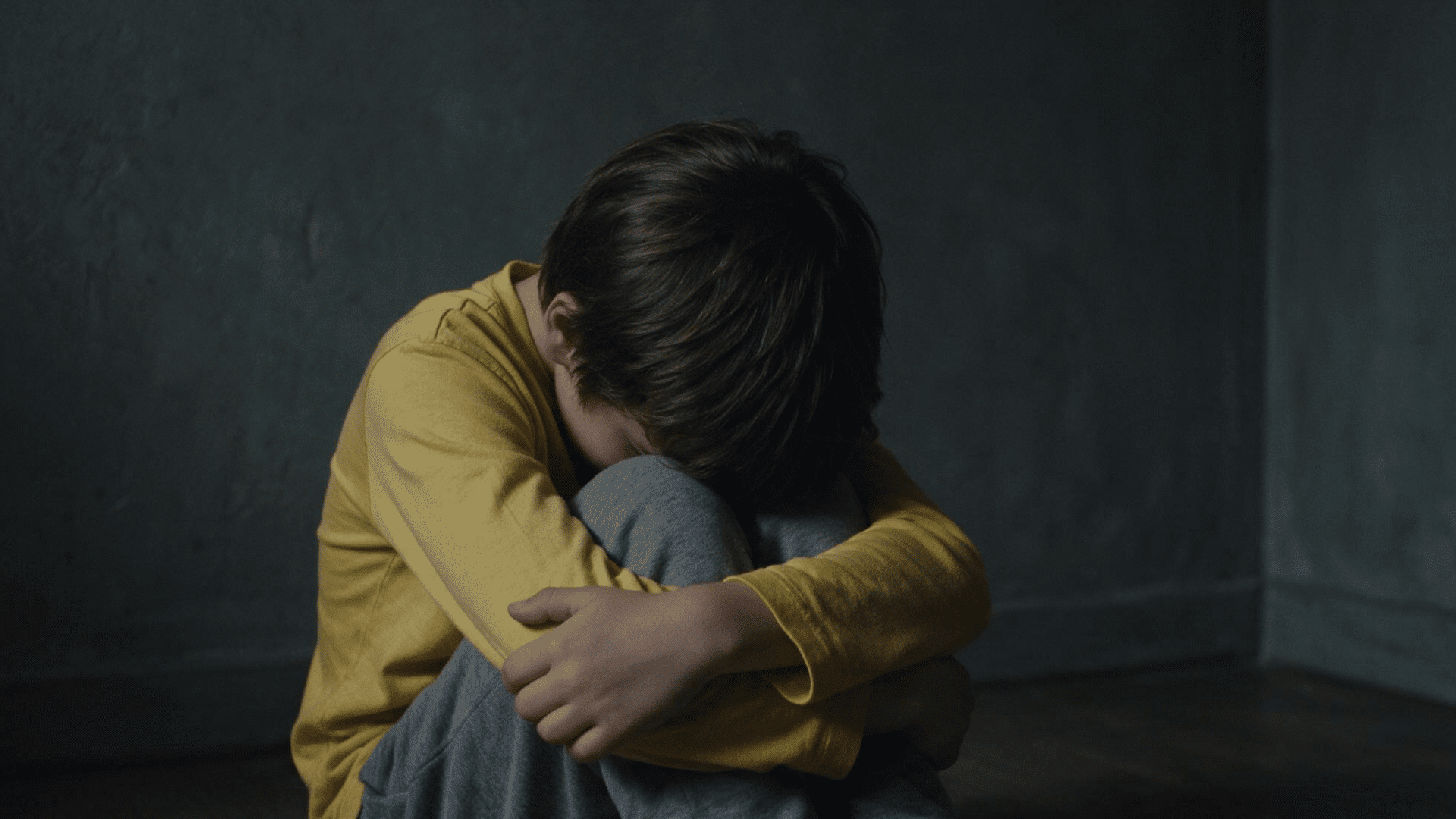 a young boy in a yellow shirt sits on the floor with his head tucked down in his knees in a dark room