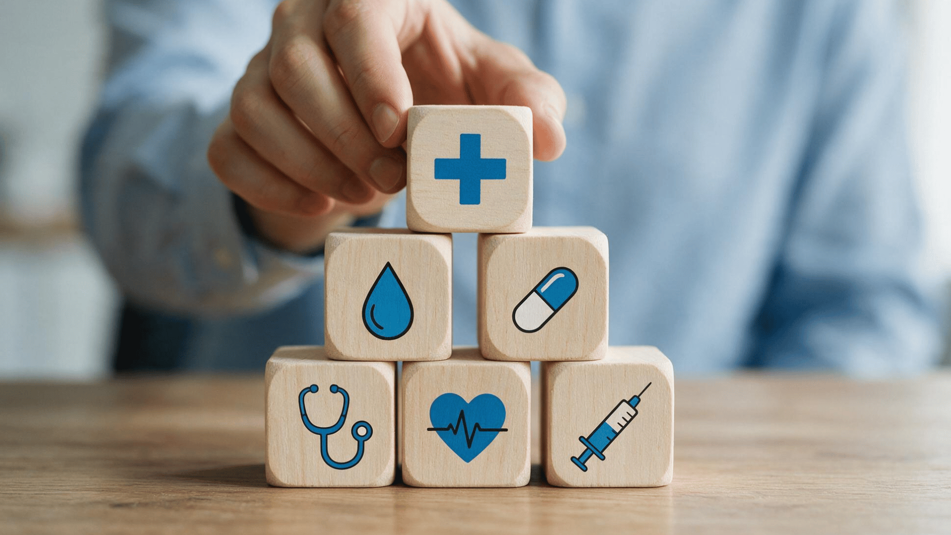 a hand places a medical cross block atop a pyramid of wooden blocks featuring healthcare icons on a desk